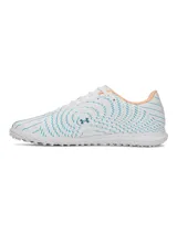 Championes de fútbol Under Armour Shadow Select 4 Turf, diseñados para césped artificial. Presentan un diseño aerodinámico en color blanco con detalles en azul petróleo y acentos en naranja. Cuentan con empeine sintético texturizado para mejor control, mediasuela Charged Cushioning para amortiguación reactiva y suela de goma con tacos multidireccionales.