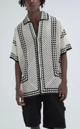 Camisa de manga corta color beige, tejida con punto abierto (símil crochet) y detalles en negro. Tiene cuello solapa, cierre frontal con botones y calce oversize.