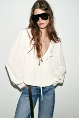 Blusa de georgette en color blanco, con escote en pico, detalle de volantes y lazada frontal. Presenta mangas largas abullonadas con puños ajustados y un bajo asimétrico con diseño tipo globo.