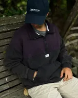 Buzo de corte oversized con medio cierre frontal, confeccionado en una combinación de colores violeta en la parte superior y negro en la parte inferior. Presenta cuello alto, puños y ruedo acanalados, y un bolsillo tipo canguro frontal con etiqueta de la marca.