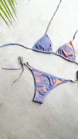 Bikini de dos piezas con estampado tie-dye en tonos violeta y naranja. El top es de triángulos movibles con tiras para atar al cuello y espalda, e incluye almohadillas desmontables. El bottom es una vedetina con tiras ajustables a los costados para atar a la medida de la cadera.