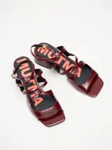 Sandalias de tacón bajo Mutma modelo Sando, confeccionadas en cuero con acabado de charol color bordó. Diseño de tiras cruzadas en el empeine, puntera cuadrada y correa ajustable en el tobillo con hebilla metálica. Tacón bloque de altura media. Plantilla con estampado de la marca en contraste.