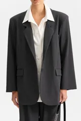 Blazer de corte amplio en color gris oscuro, con solapas de muesca y bolsillos de ojal. La prenda simula llevar una camisa blanca de cuello ancho por debajo, con los botones visibles en el frente.