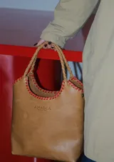 Bolso tipo matera de cuero color suela, con costuras decorativas en hilo rojo en los bordes y manijas trenzadas. Presenta el logo de la marca grabado en el frente.