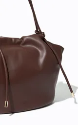 Cartera tipo tote de gran tamaño color chocolate, con diseño flexible y cierre de cordón ajustable. Presenta un acabado liso y minimalista con el logo de la marca grabado en la parte frontal.