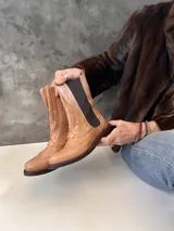 Bota tipo borceguí de cuero color beige, con diseño estilo brogue y elástico lateral. Cuenta con cierre interno y suela de goma con taco bajo.