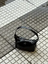 Bolso de hombro negro de charol, con forma alargada y cierre principal con cremallera. Presenta un dije metálico con forma de estrella de cuatro puntas en el frente y detalles de cremalleras decorativas en los laterales.