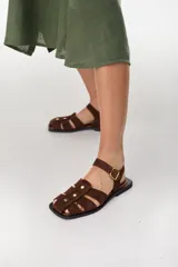Sandalias planas de gamuza marrón, estilo pescador, con tiras entrelazadas, tachas doradas y tira ajustable con hebilla en el tobillo.