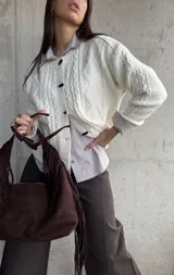 Cardigan de punto trenzado color natural, con cuello redondo, cierre frontal de botones negros y mangas largas con puños acanalados.