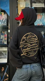 Canguro negro de corte oversize con capucha y bolsillo frontal. Presenta un diseño gráfico circular estampado en color beige en la espalda con tipografía estilizada y detalles de texto en la parte inferior.