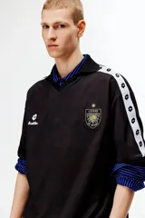 Camiseta de futbol de manga corta con corte holgado, color negro con estampa sutil y bandas blancas con rombos en las mangas. Presenta cuello en V con solapa y esc11udo bordado en el pecho.