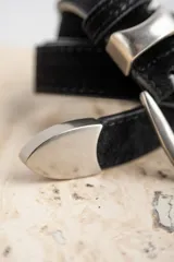 Cinto de cuero negro con textura de gamuza, presenta una hebilla metálica plateada de diseño clásico y puntera metálica a juego.