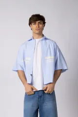 Camisa de manga corta de corte boxy, confeccionada en popelina a rayas celestes y blancas. Presenta cuello camisero clásico, cierre frontal con botones y estampado tipográfico en el pecho.