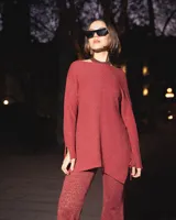 Blusa de tejido de punto color rojo oscuro con hilos dorados brillantes, manga larga y cuello tipo choker. Presenta un corte asimétrico y aberturas en los puños.