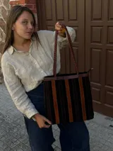 Cartera tipo tote de cuero con diseño de franjas verticales multicolores en tonos tierra, rojo y azul. Cuenta con correas trenzadas de cuero y cierre superior.
