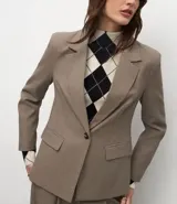 Blazer de sastrería color marrón, con solapas clásicas, cierre frontal de un botón, bolsillos con solapa y mangas largas. Presenta un corte entallado que define la silueta.