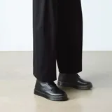 Botas de caña media Dr. Martens modelo Brookline, confeccionadas en cuero negro con acabado ceroso. Presentan cierre de cordones con seis ojales, suela gruesa con costuras amarillas visibles y tirador característico en el talón.