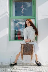 Cartera de mano tipo bolso con flecos laterales, confeccionada en cuero con acabado texturizado en tono claro. Presenta asas circulares metálicas, correa larga ajustable y extraíble, y un diseño de estilo boho.