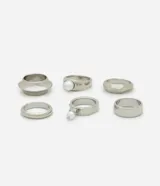 Set de seis anillos de metal plateado con diseños variados, incluyendo bandas lisas, texturizadas y modelos con aplicaciones de perlas sintéticas.
