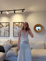 Vestido largo blanco con escote en V, decorado con flecos cortos en el busto y la cadera. Detalle de lentejuelas plateadas en el busto y la cadera. Breteles metalizados cruzados en la espalda.