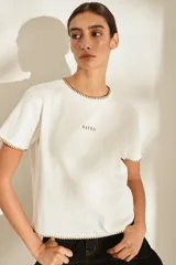 Remera marrón de manga corta y cuello redondo, con detalles de costuras decorativas en contraste color crudo en el cuello, los puños y el dobladillo inferior. Cuenta con el nombre de la marca impreso en el pecho.