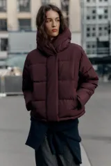 Campera puffer acolchada de color negro, con cuello envolvente, capucha integrada, manga larga, bolsillos laterales ocultos y bajo con elástico. Cierre frontal con botones a presión ocultos por solapa.