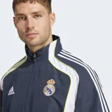 Campera deportiva Adidas de entrenamiento del Real Madrid, color azul marino con paneles laterales blancos y ribetes finos en amarillo neón. Presenta cierre frontal, cuello alto y el escudo del club bordado en el pecho.