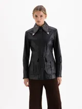 Parka de cuero vacuno color negro, con diseño utilitario que incluye bolsillos frontales con tapa y fuelle. Presenta un cuello con detalle de tachas metálicas y cierres en los puños.