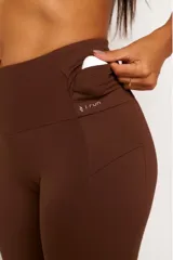 Calza deportiva de tiro alto color marrón, confeccionada en tejido técnico de alto rendimiento con compresión. Presenta bolsillos laterales funcionales y detalles reflectivos para mayor visibilidad.