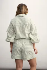 Camisa a rayas verticales color beige y blanco, con cuello clásico, cierre frontal con botón dorado, mangas largas abullonadas con puños elásticos, dos bolsillos frontales y corte holgado.