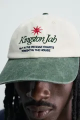 Gorra de seis paneles con efecto lavado, de color blanco crudo con visera en verde militar. Presenta un bordado frontal contrastante con texto en negro y una estrella roja.
