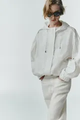 Chaqueta blanca de cuello redondo con capucha ajustable mediante cordones elásticos. Presenta manga larga con puños elásticos, bolsillos delanteros y bajo asimétrico ajustable con cordones y stoppers. El cierre frontal combina cremallera y botones ocultos bajo una solapa.