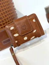 Mini billetera de cuero con diseño de pelo animal print en tonos marrón y blanco. Presenta un cierre de solapa con broche y un logo de búho grabado en el frente, además de una correa de muñeca extraíble.