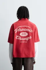 Camisa de algodón con estructura canvas, color rojo lavado, de corte relajado (relaxed fit) y manga corta. Posee cuello solapa, dos bolsillos de plastrón en el pecho y cierre frontal de botonadura. Presenta bordado y estampado de texto a contraste en el delantero.