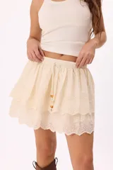Minifalda color crema con bordado calado tipo broderie, cintura elastizada con cordón ajustable y detalle de cuentas de madera. Diseño de doble capa con volados.