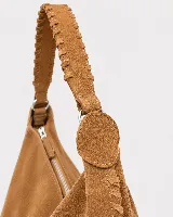 Bolso tipo hobo de gran tamaño, confeccionado en gamuza negra con detalles de costuras artesanales y tientos decorativos en los bordes. Cuenta con un asa trenzada con pespuntes a mano y cierre superior.