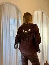Campera marrón de gamuza con flecos en la espalda y las mangas, solapas anchas y detalles de pines metálicos en forma de estrella y figuras abstractas.