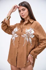Camisa marrón de manga larga con puños anchos, cuello camisero y botones frontales. Presenta bordados florales en blanco con lentejuelas y mostacillas en la parte delantera.