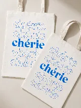 Bolsa de tela tipo tote bag color blanco crudo, con estampado tipográfico repetido con la palabra "chérie" en tonos azules. Cuenta con asas dobles de tela para llevar al hombro.