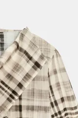 Camisa de manga larga con estampado de cuadros en tonos neutros. Presenta un diseño con cuello tipo pañuelo, cierre frontal cruzado con botones y un corte asimétrico en el ruedo.