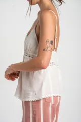 Top blanco de algodón peinado con tirantes finos, escote en V con detalle de encaje y cierre frontal abotonado. Presenta inserciones de encaje transparente en el abdomen y un diseño fluido en la parte inferior.