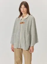 Capa de tejido de punto en color gris melange, con diseño abierto y cierre frontal mediante un botón tipo duffle de madera con detalle en cuero. Presenta bolsillos laterales amplios y bordes con costuras a la vista.