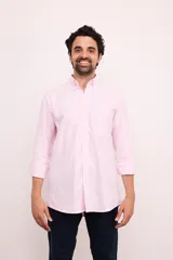 Camisa de corte clásico confeccionada en algodón, con diseño de rayas verticales finas en color rosa sobre fondo blanco. Presenta cuello abotonado, cierre frontal con botones, mangas largas con puños abotonados y un bolsillo en el pecho con el logo de la marca bordado.