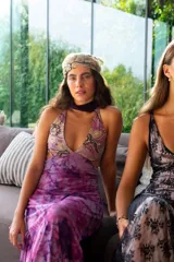 Vestido largo con estampado tie-dye en tonos violeta y fucsia, escote en V con detalles bordados dorados, cut-outs laterales bajo el busto y tirantes finos que se cruzan y atan en la espalda.