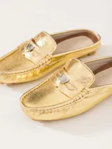 Mocasín tipo mule (destalonado) de cuero vacuno flor con textura croco, color dorado metalizado. Presenta un detalle metálico en forma de corazón sobre el empeine y suela de goma con relieve.