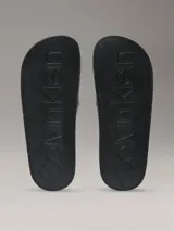 Ojotas Calvin Klein con diseño de monograma de la marca en color beige sobre fondo negro. Presentan una suela anatómica contorneada para mayor confort y una tira superior de material sintético.