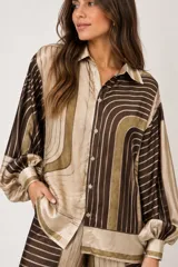 Camisa de seda estampada con diseño geométrico de rayas y líneas curvas en tonos marrón, verde salvia y beige, con cuello camisero, cierre frontal con botones y mangas largas abullonadas con puños ajustados.