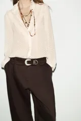 Camisa de cuello redondo y manga larga con puños, confeccionada en tejido semitransparente con textura de pequeños puntos en relieve. Presenta volantes decorativos en el frente y cierre frontal con botones ocultos.