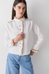 Camisa de manga larga en color blanco, confeccionada en tejido de lino, con detalles de volados en crochet en el cuello, la botonadura frontal y los puños. Presenta detalles de costuras en color ocre en los hombros y puños, con cierre frontal de botones.