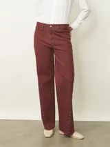 Jean de corte recto y tiro medio en color bordeaux, con detalle de tachas metálicas a lo largo de las costuras laterales y ruedo desflecado.